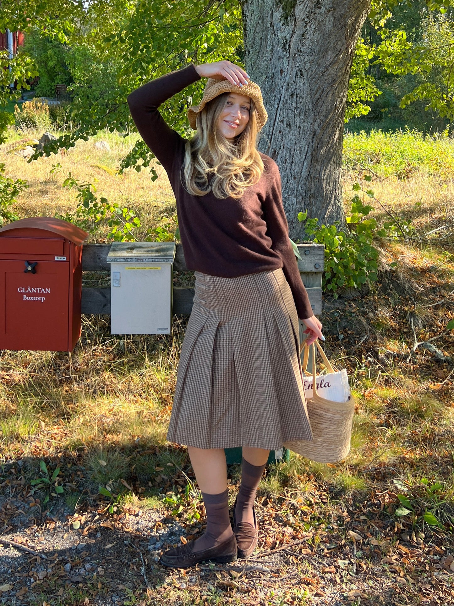 Woman in a brown sweater and skirt standing outdoors near a tree and mailbox.