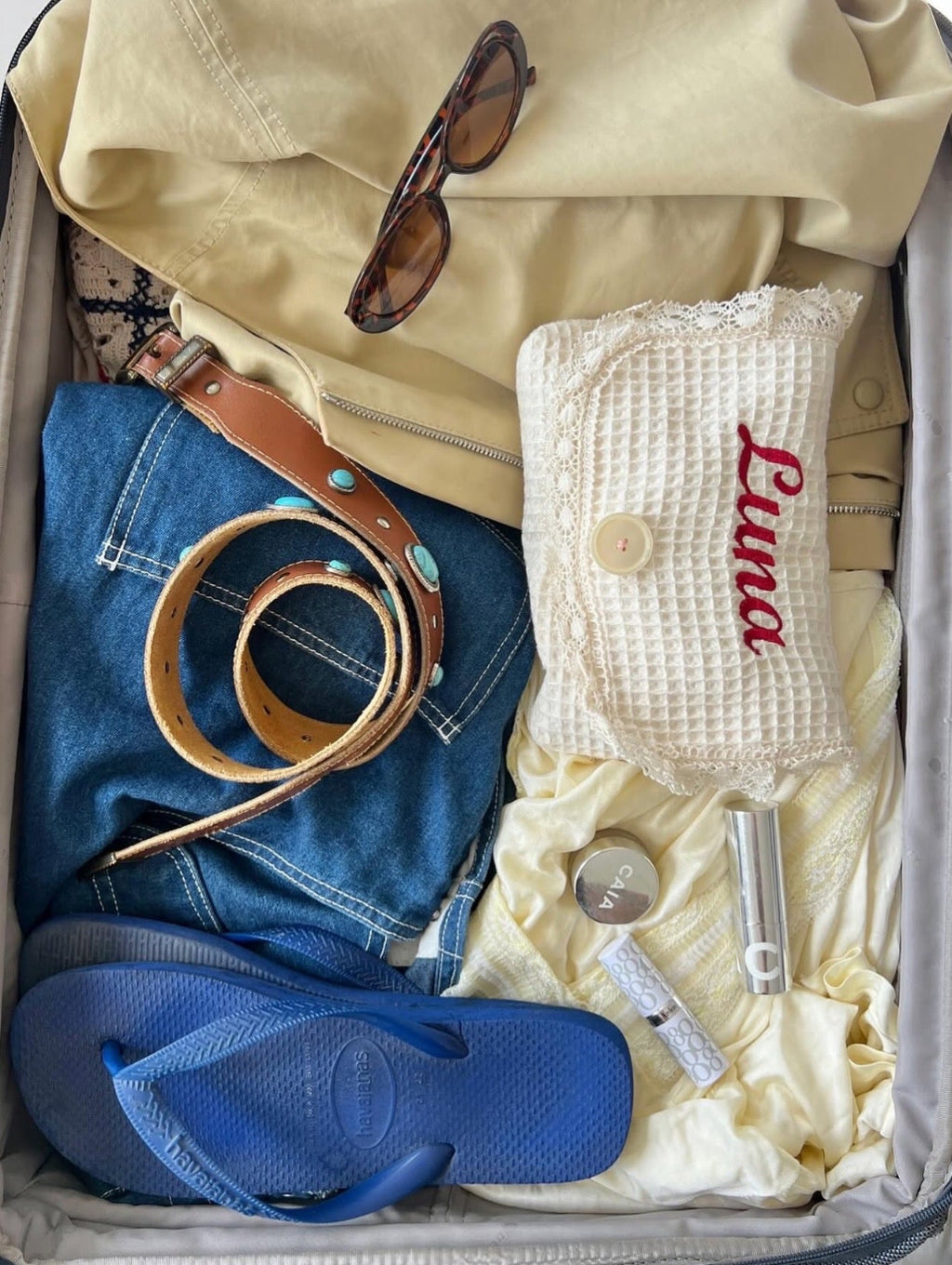 Open suitcase with clothes, shoes, and accessories including a yellow jacket, blue jeans, blue flip-flops, and sunglasses.