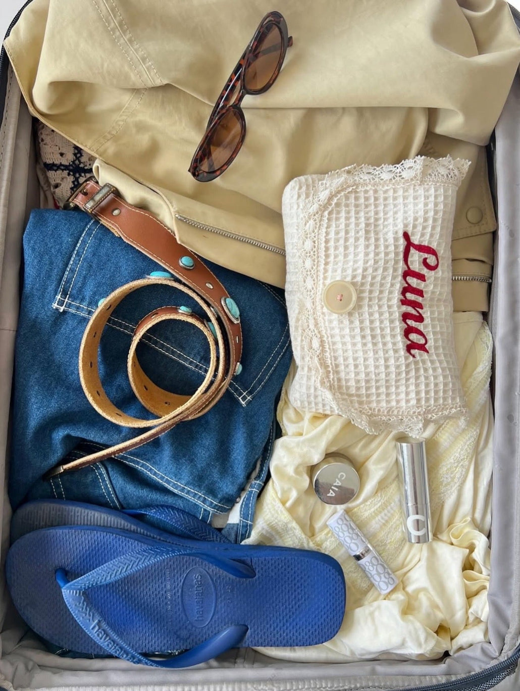 Open suitcase with clothes, shoes, and accessories including a yellow jacket, blue jeans, blue flip-flops, and sunglasses.